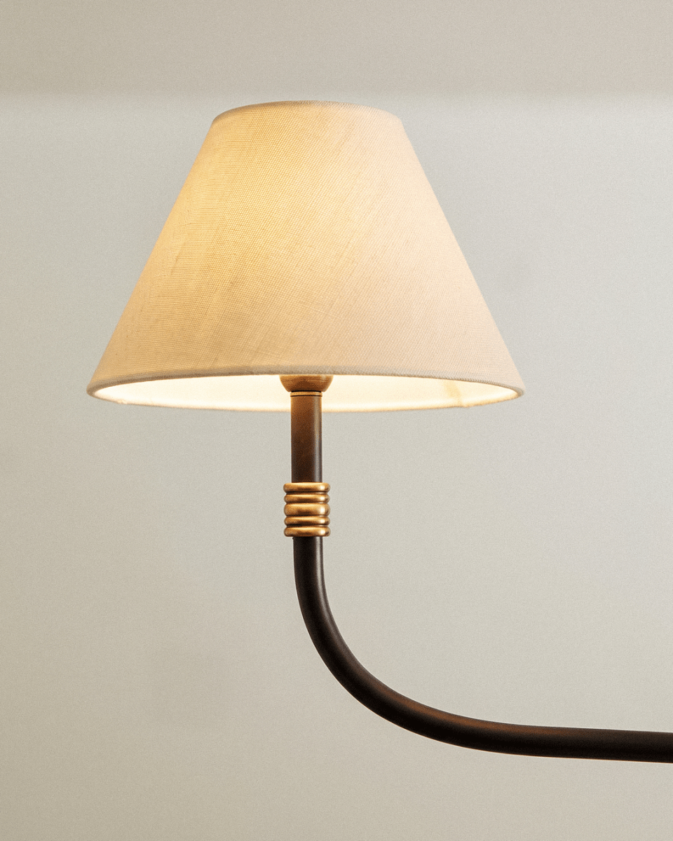 Close view of a Clérachic chandelier arm, with a cream linen shade, ribbed brass collar and curved black iron stem.