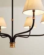 Close detail of the Clérachic chandelier hub, where black iron arms meet a brass collar beneath cream linen shades.