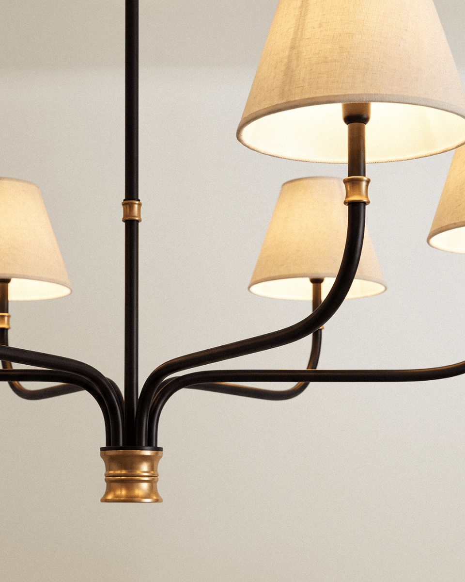 Close detail of the Clérachic chandelier hub, where black iron arms meet a brass collar beneath cream linen shades.