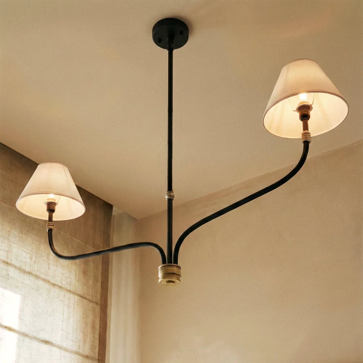 Clérachic two-arm chandelier seen from below, its cream shades lit warm against a pale ceiling and linen window blind.