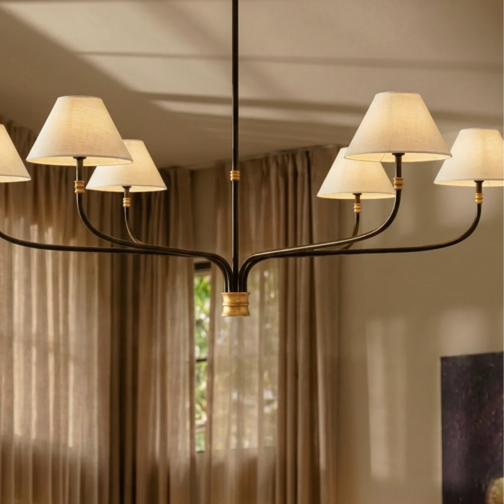Clérachic six-arm chandelier with cream linen shades glowing warm in front of sheer curtains and a window.