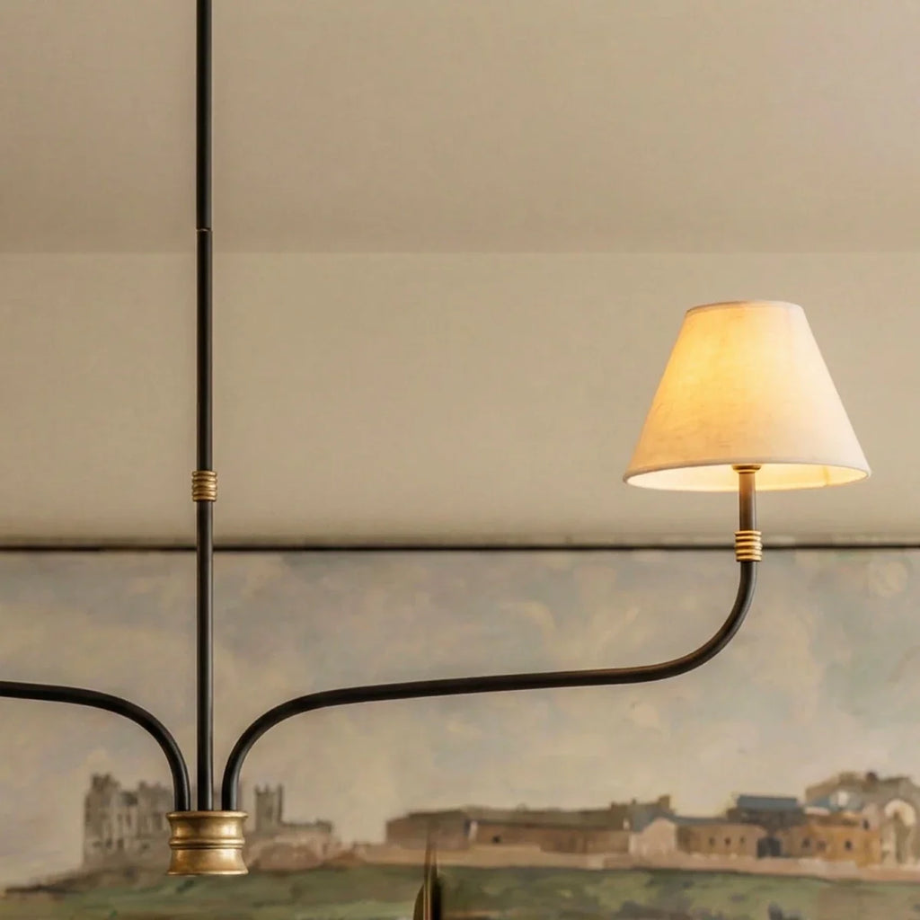 Clérachic chandelier arm with a lit cream shade, ribbed brass collar and black iron stem, set against a landscape painting.
