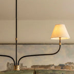 Clérachic chandelier arm with a lit cream shade, ribbed brass collar and black iron stem, set against a landscape painting.