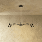 A matte black iron chandelier with slender branching arms and cylindrical heads hangs against a plaster wall, Clérachic hero shot.