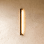 The black column wall light glows warm against plaster in this closer Clérachic hero shot, its slim vertical profile catching soft shadow.