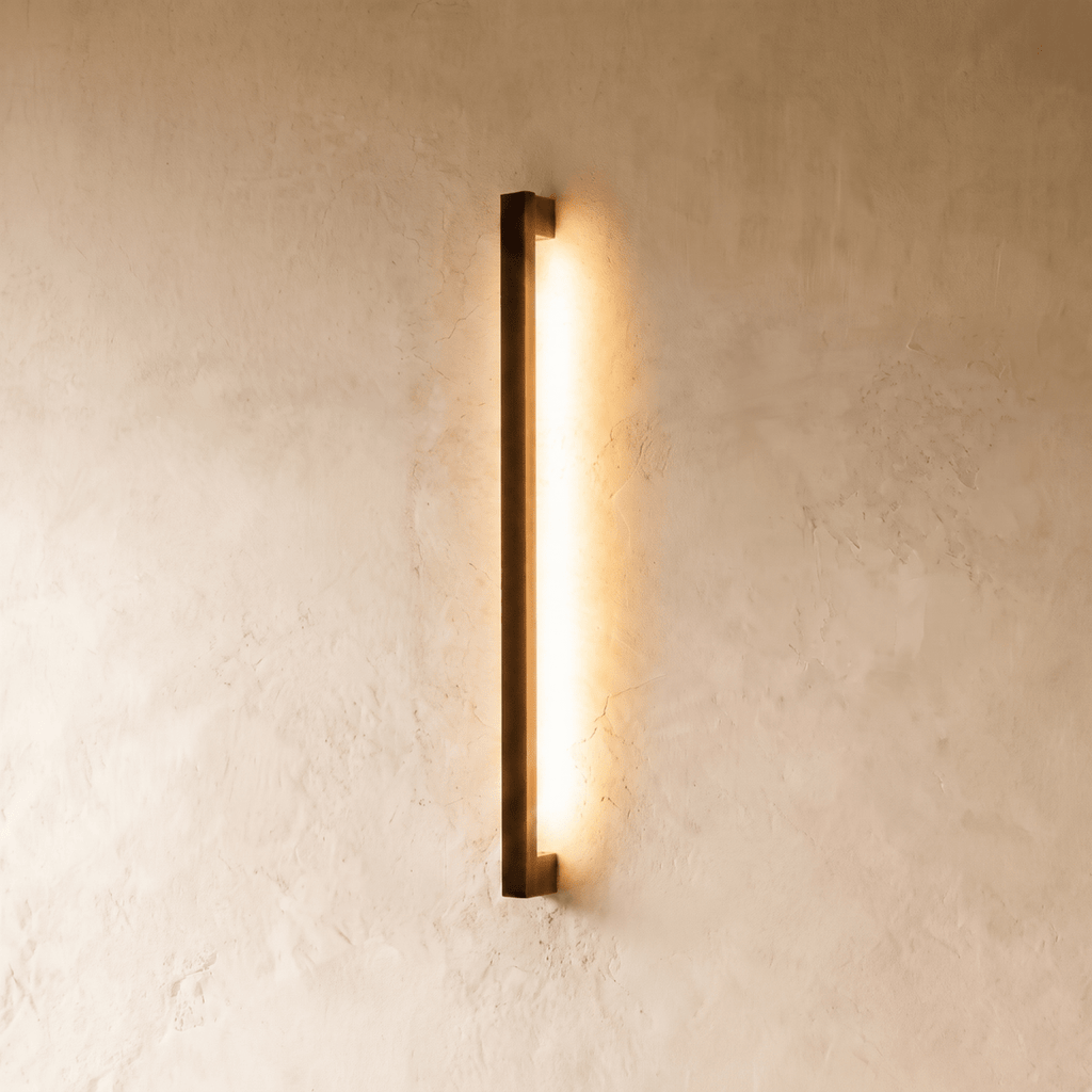 A further lit hero shot of the slim black column wall light shows its even warm wash down a textured plaster wall, styled by Clérachic.