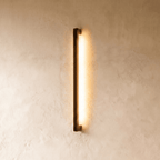 A further lit hero shot of the slim black column wall light shows its even warm wash down a textured plaster wall, styled by Clérachic.