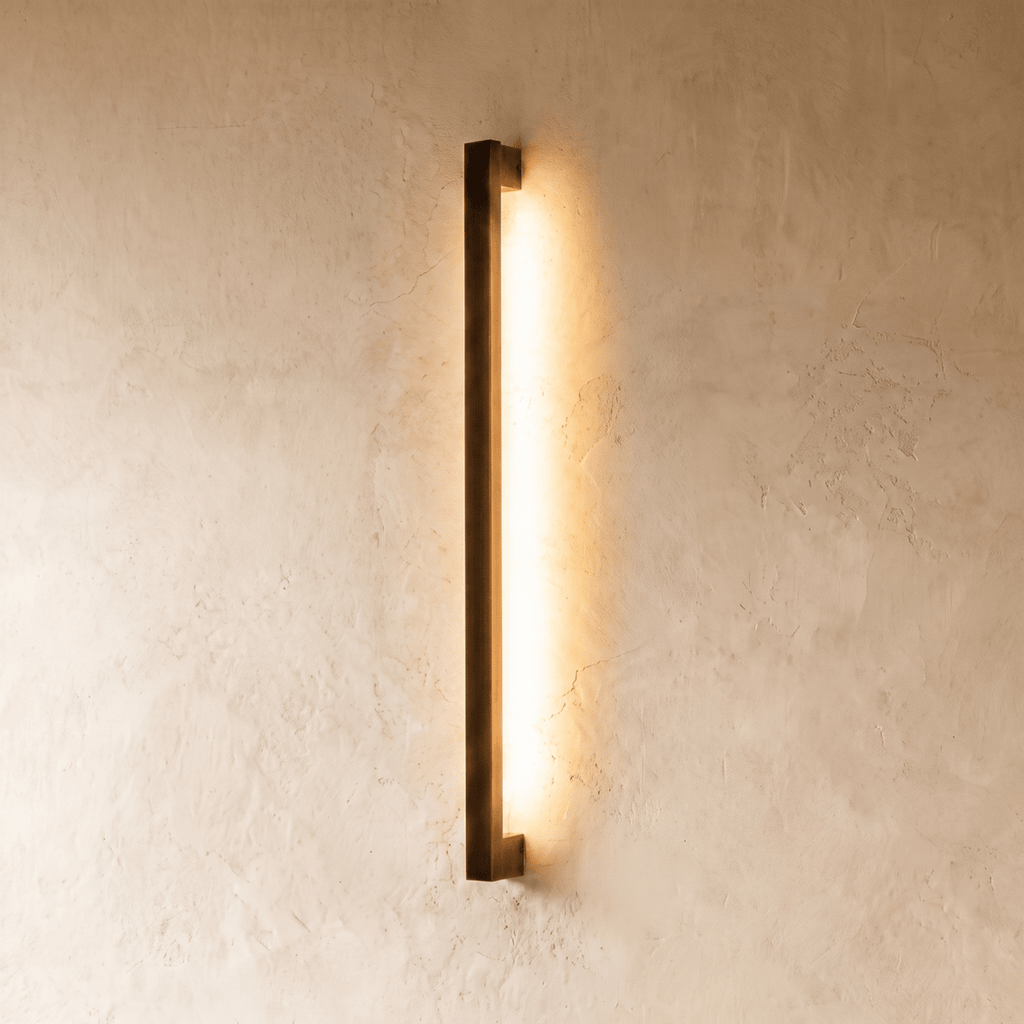 A darker matte-black column wall light glows warm against plaster in this Clérachic hero shot, its clean square profile visible in shadow.