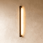 A darker matte-black column wall light glows warm against plaster in this Clérachic hero shot, its clean square profile visible in shadow.