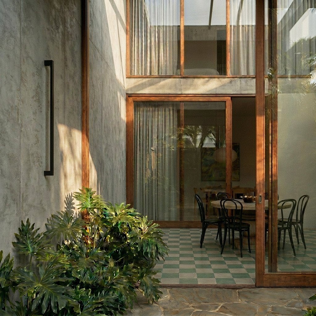 The slim black column wall light sits unlit on a concrete wall beside a stone accent and trailing greenery, Clérachic lifestyle shot.