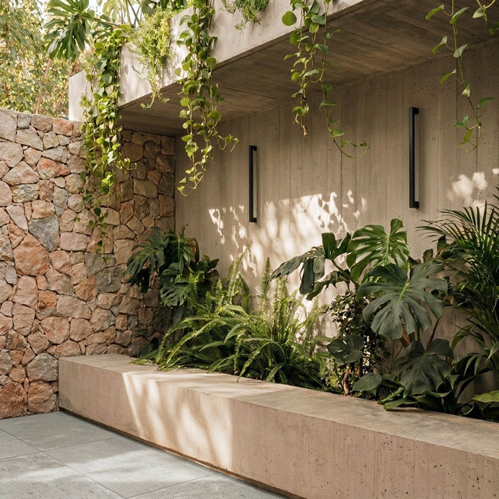 Two black column wall lights flank a concrete slat wall, unlit by day, framed by hanging vines and ferns in this Clérachic lifestyle shot.