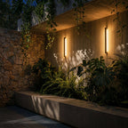 At dusk, two black column wall lights cast a warm glow down a stone courtyard wall amid trailing plants, Clérachic lifestyle shot.