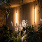 A tighter night view shows the pair of black column wall lights glowing warm against the stone courtyard wall, Clérachic lifestyle shot.
