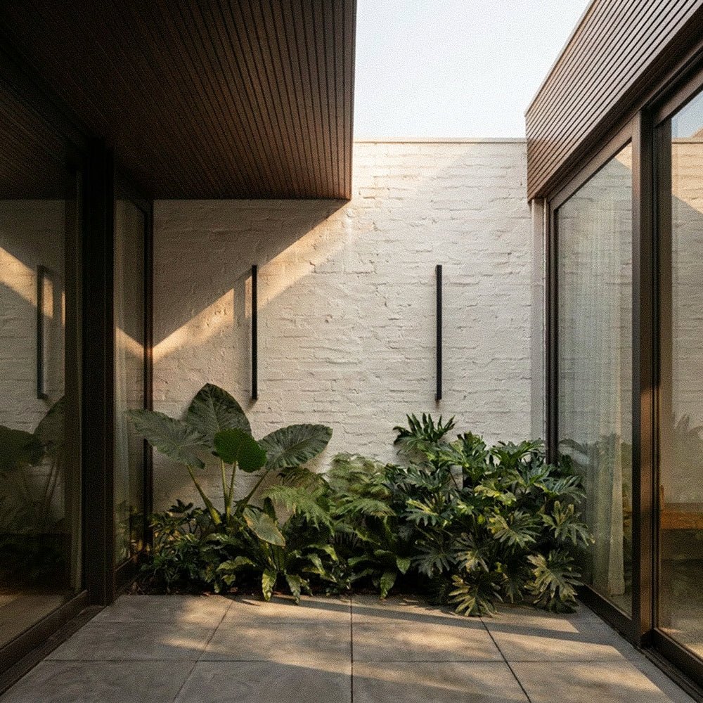 Two black column wall lights stand unlit against a whitewashed brick wall between glazed doors and leafy planting, Clérachic lifestyle shot.