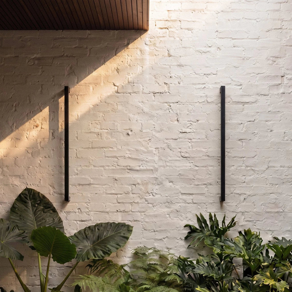 A close-up on two black column wall lights mounted on whitewashed brick, framed by broad tropical leaves, Clérachic detail shot.