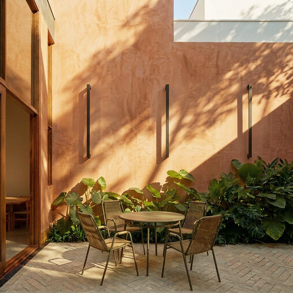 Three black column wall lights line a sun-warmed terracotta courtyard wall above a rattan table and chairs in this Clérachic lifestyle shot.