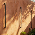 A closer view of the black column wall lights against terracotta stucco shows dappled tree shadows across the wall, Clérachic detail shot.