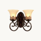 A two-arm Clérachic wall lamp with scrolled dark bronze arms and cream glass bell shades, lit on a white backdrop.