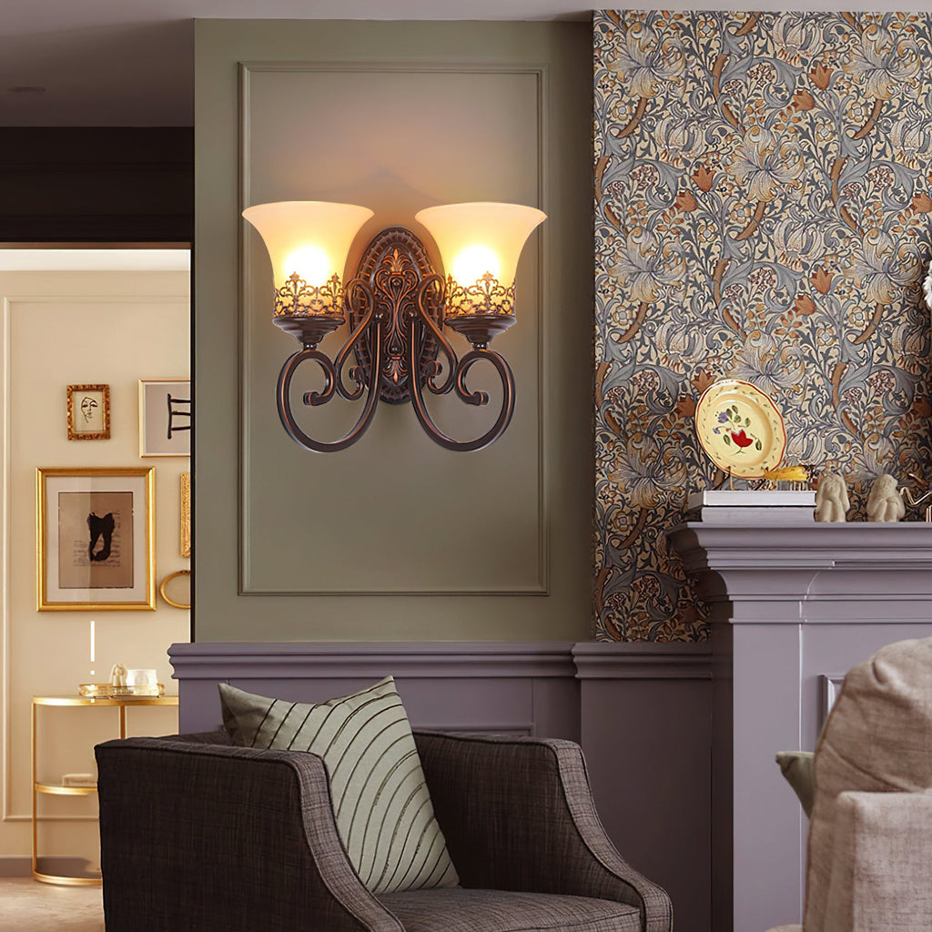 A two-arm Clérachic wall lamp glows on a sage panel beside patterned wallpaper, above a striped cushion on an armchair.