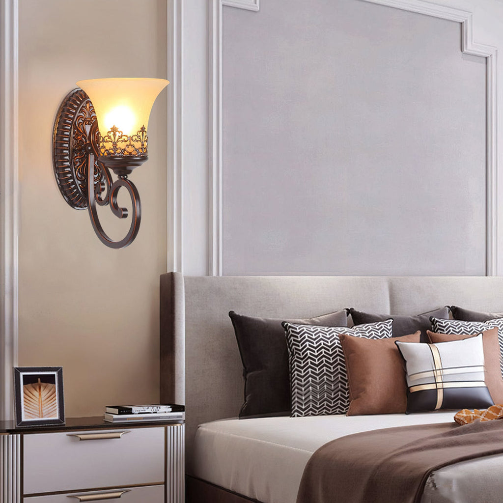 A single-arm Clérachic wall lamp glows above a bedside table, by a bed dressed in patterned and brown cushions.