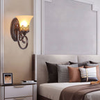 A single-arm Clérachic wall lamp glows above a bedside table, by a bed dressed in patterned and brown cushions.