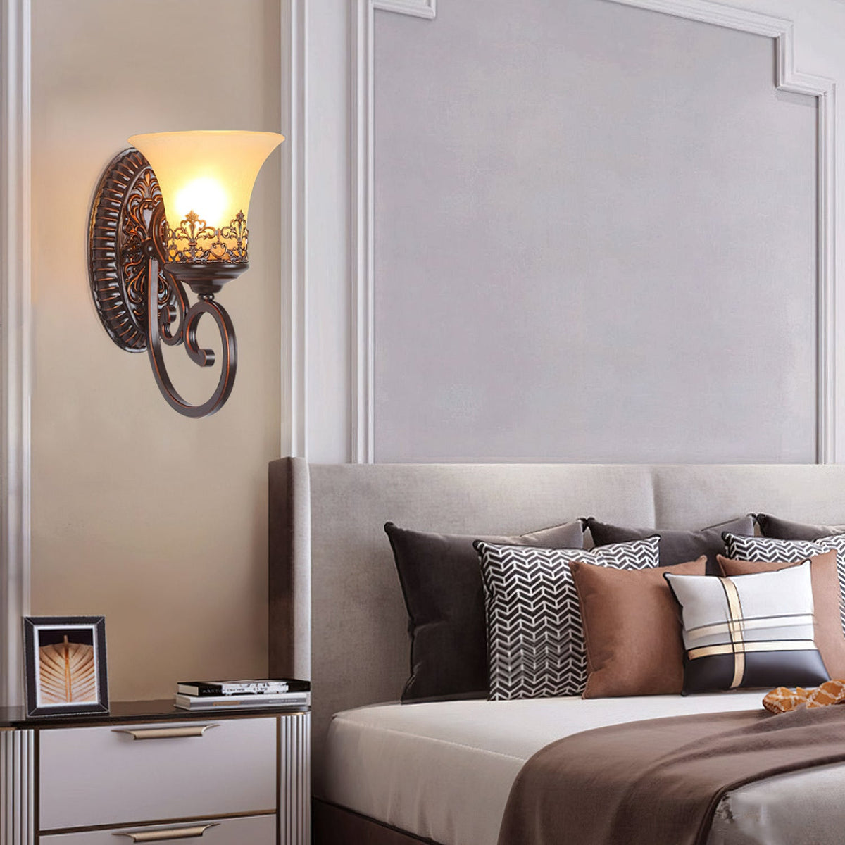 A single-arm Clérachic wall lamp glows above a bedside table, by a bed dressed in patterned and brown cushions.