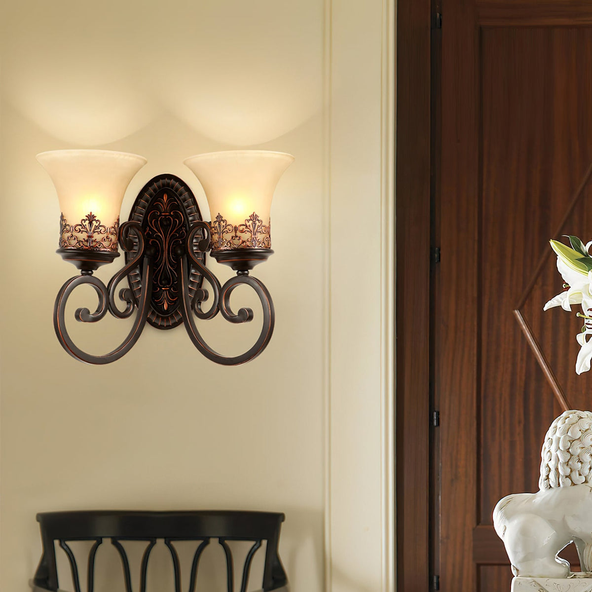 A two-arm Clérachic wall lamp lights a cream wall beside a dark wood door, with a black chair back below.