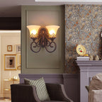 A two-arm Clérachic wall lamp glows against a sage panel, with a gilt plate on the mantel and an armchair below.