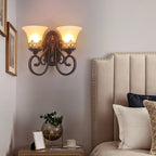 A two-arm Clérachic wall lamp glows above a bedside table, with a cream headboard and navy and white cushions.