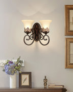 A two-arm Clérachic wall lamp glows on a grey wall between gilt-framed pictures, above a vase of flowers and a small framed print.
