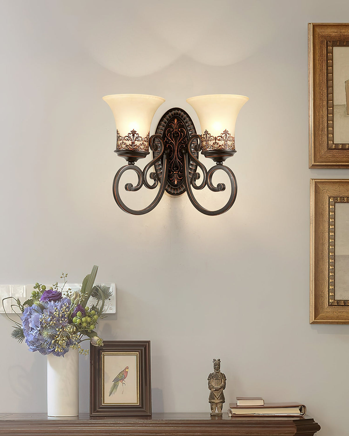A two-arm Clérachic wall lamp glows on a grey wall between gilt-framed pictures, above a vase of flowers and a small framed print.