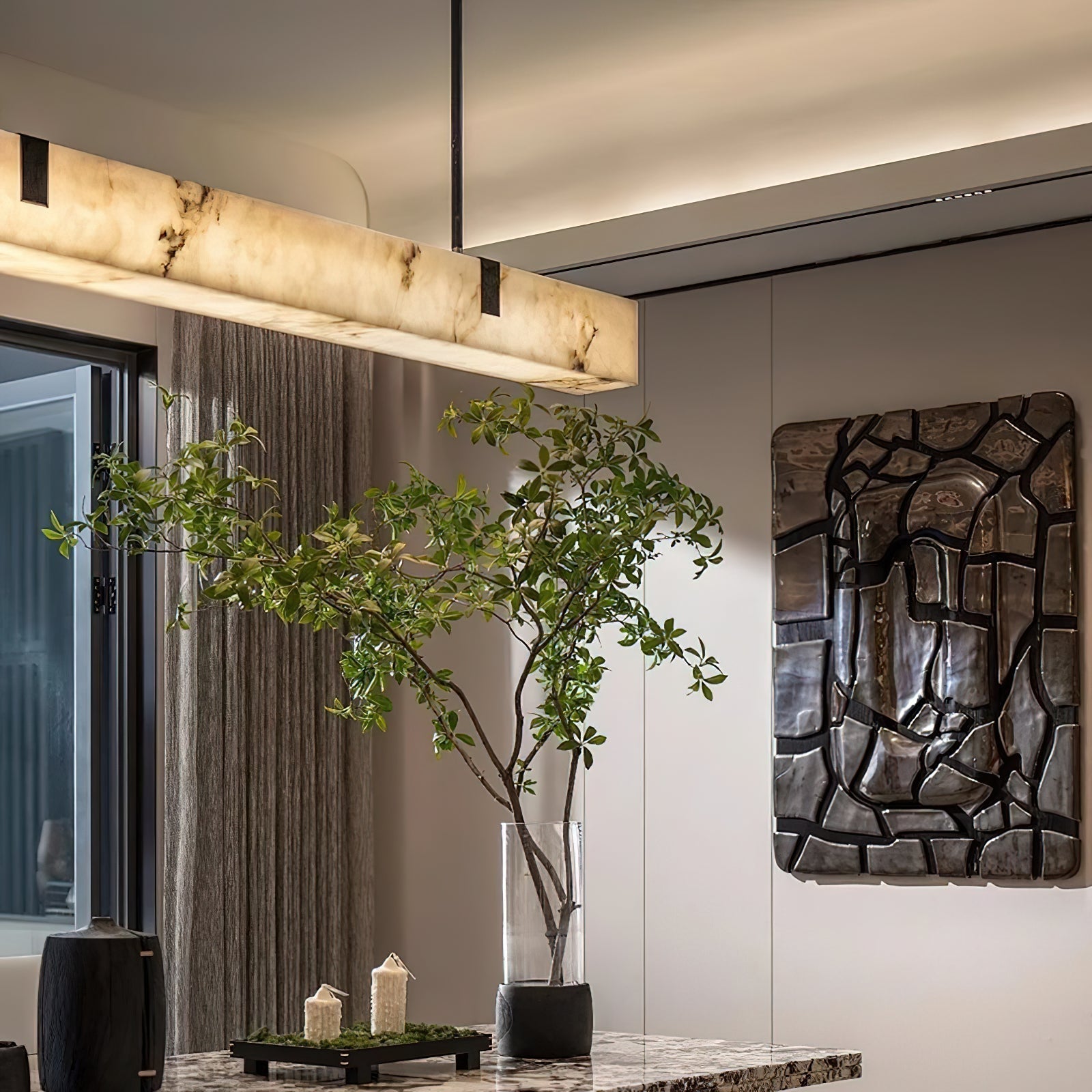 The Clérachic Black Linear Chandelier glows warm above a marble dining table beside a potted olive tree and dark wall art.