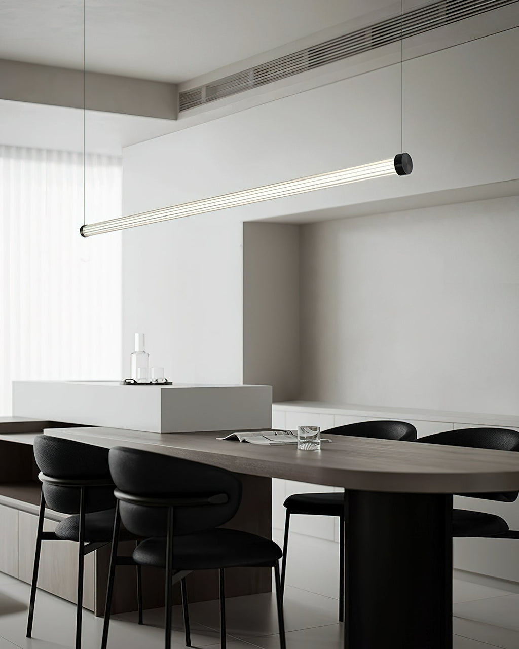 Tilted view of a Clérachic linear pendant above a dark oval table, with a glass carafe on a pale island behind it.
