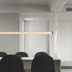 Close crop of a Clérachic linear pendant running across the frame, above black rounded dining chairs in a pale open-plan room.