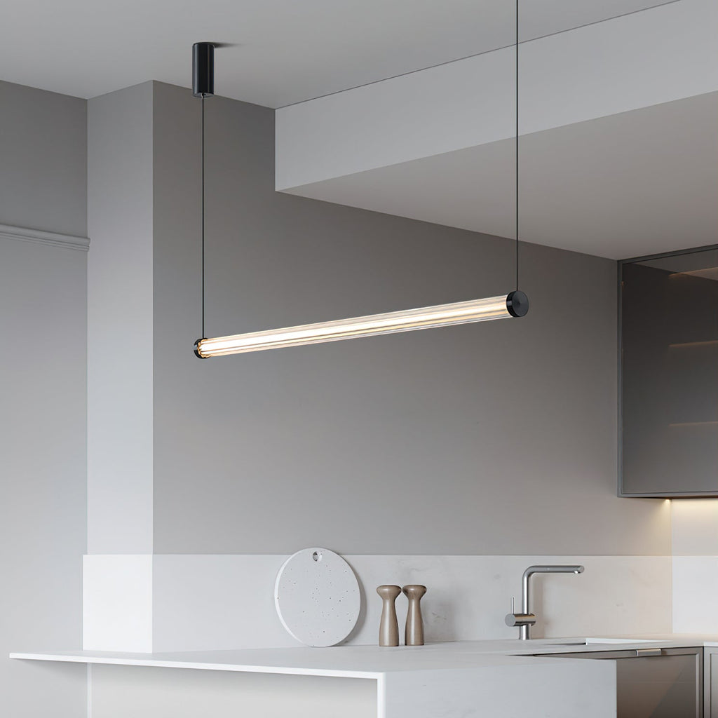 This Clérachic linear pendant hangs at a slant in a white kitchen, with a marble counter, a tap and wooden pepper mills below.