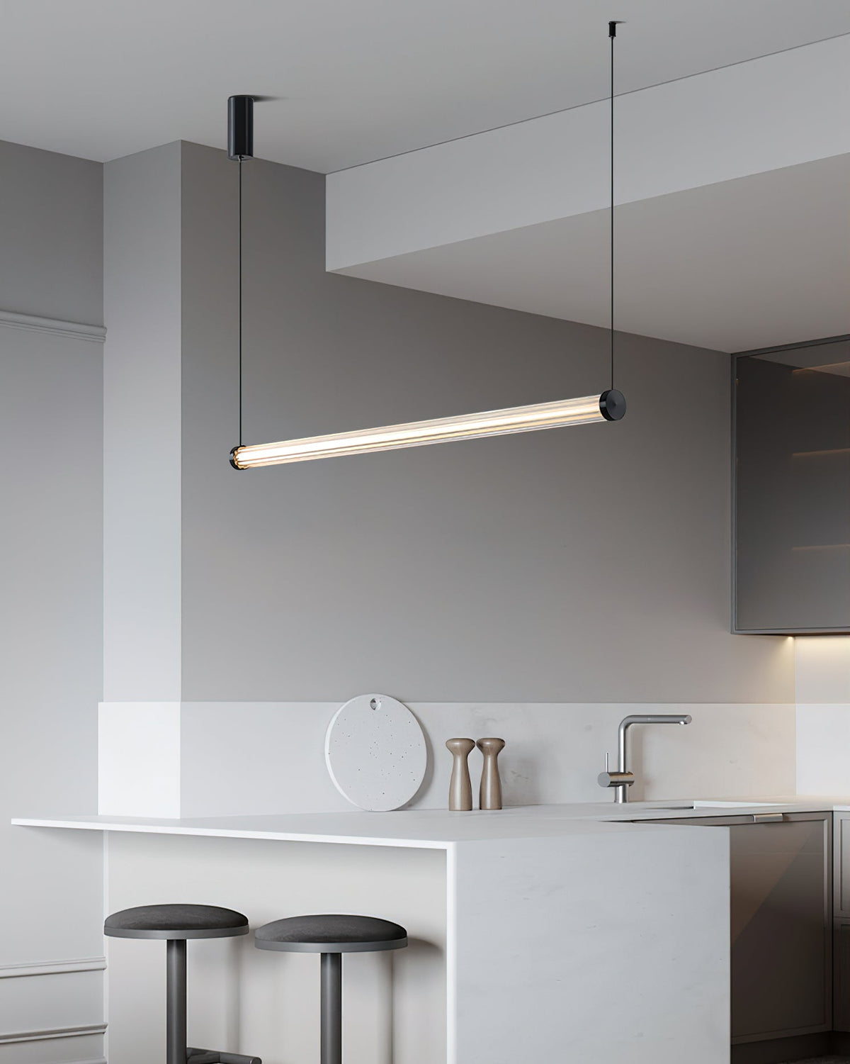 A Clérachic linear pendant tilted above a white kitchen island with two dark stools, a marble counter and a round board behind.