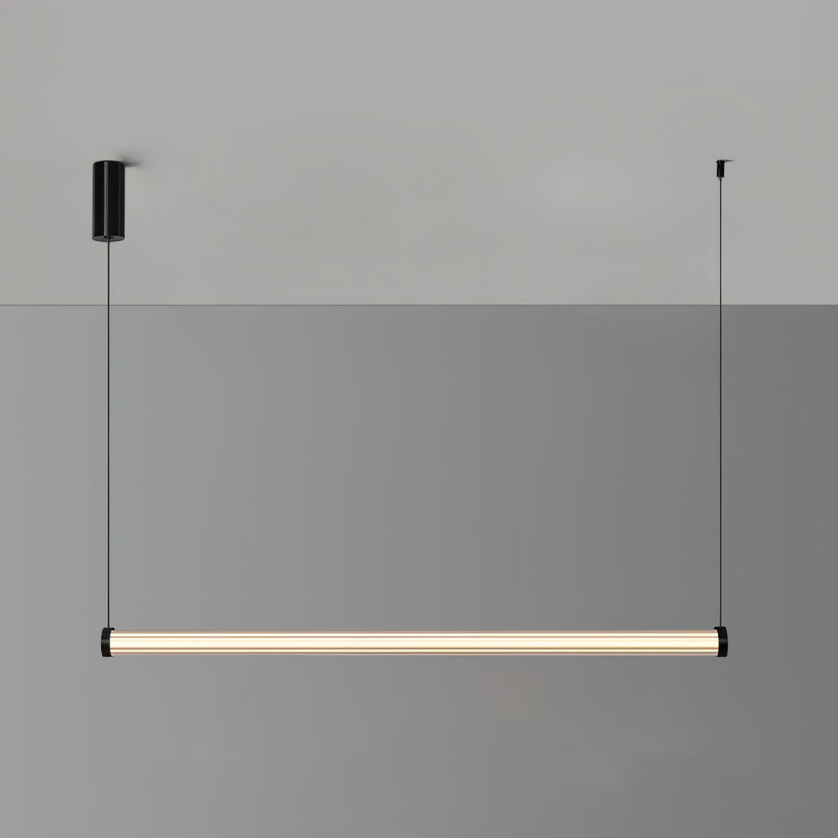 A level Clérachic linear pendant lit warm against a two-tone grey wall, with its black canopy and cable at either end.