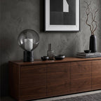 On a walnut sideboard under a framed black and white print, this Clérachic table lamp sits beside dark bowls and a tall black vase.