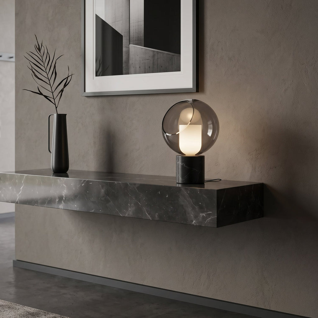 Lit on a floating stone shelf against a grey plaster wall, this Clérachic table lamp stands next to a slim black vase with dried leaves.