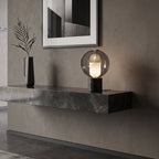 Lit on a floating stone shelf against a grey plaster wall, this Clérachic table lamp stands next to a slim black vase with dried leaves.