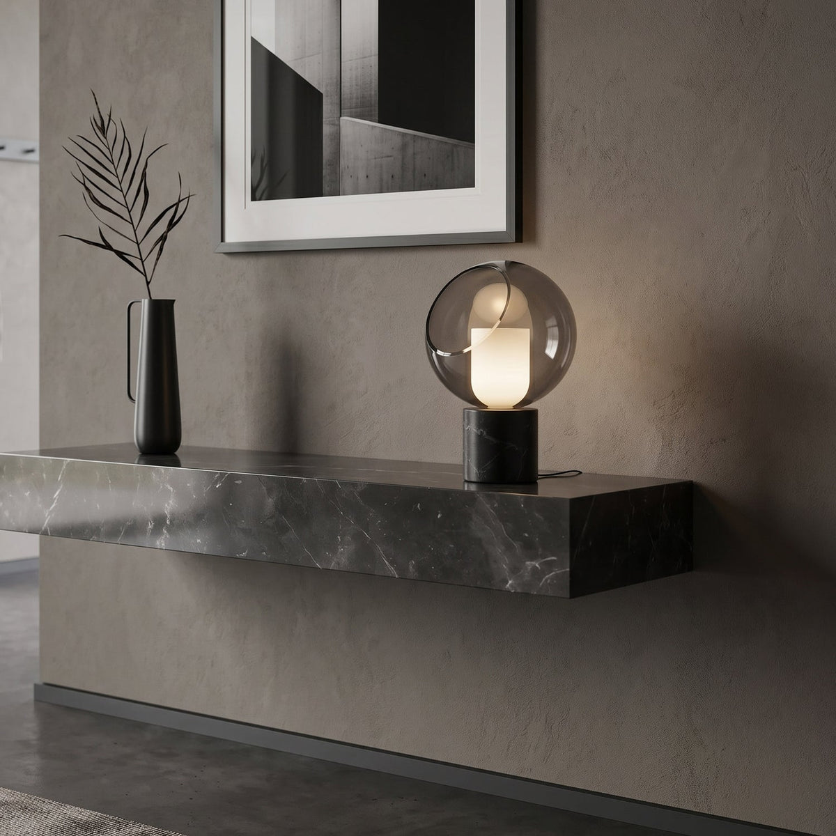 Lit on a floating stone shelf against a grey plaster wall, this Clérachic table lamp stands next to a slim black vase with dried leaves.