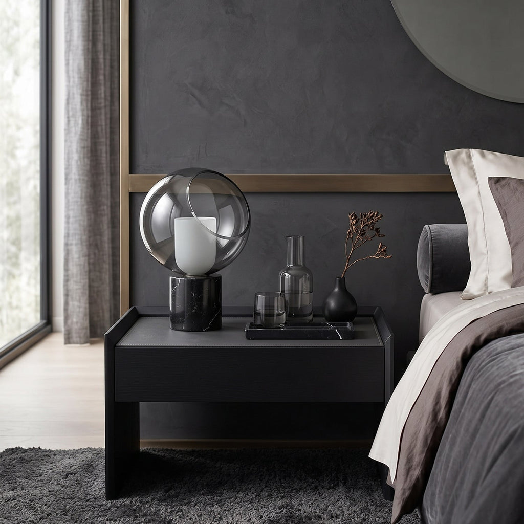 On a black bedside table next to a grey bed, this Clérachic table lamp glows beside a glass carafe and a small vase.