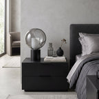 This Clérachic table lamp sits on a two-drawer black nightstand, with a stack of books, a glass carafe and a grey headboard behind it.