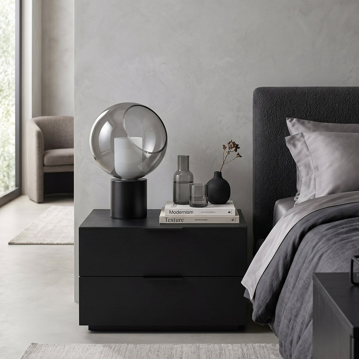 This Clérachic table lamp sits on a two-drawer black nightstand, with a stack of books, a glass carafe and a grey headboard behind it.
