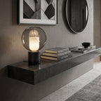 On a black stone floating shelf with books and a round mirror beside it, this Clérachic table lamp glows against a grey wall.