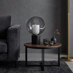 Beside a velvet sofa and a brass floor lamp, this Clérachic table lamp stands on a round wooden side table with small glass tumblers.
