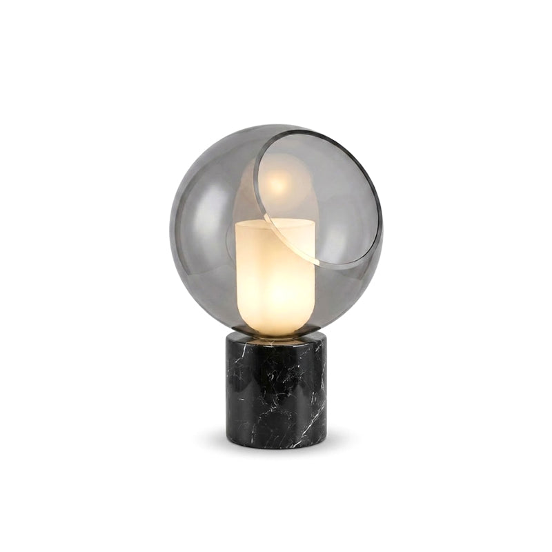 A Clérachic table lamp cut out on white, the white cylinder lit inside a smoke-grey glass globe on a black marble base.
