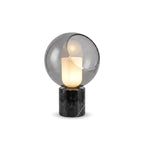 A Clérachic table lamp cut out on white, the white cylinder lit inside a smoke-grey glass globe on a black marble base.