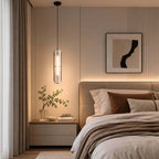 Clérachic capsule pendant lit above a bedside table, beside a bed with rust and taupe cushions and a framed abstract print.