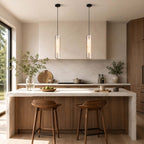 Two Clérachic capsule pendants lit over a marble kitchen island with wooden stools, in front of a tall black-framed window.